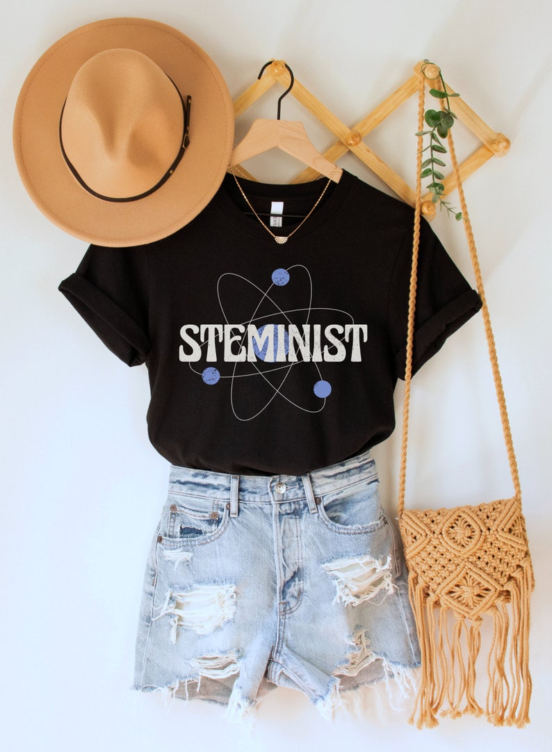 May include: Black t-shirt with a white atom graphic and the text "STEMINIST" in white letters. The shirt is on a hanger with a brown hat and a woven bag.