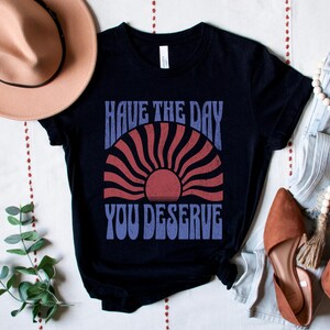 A black unisex short sleeve crew neck t-shirt that says "have the day you deserve" on the front in distressed blue retro capitalized font around a distressed image of a red wavy retro sunset