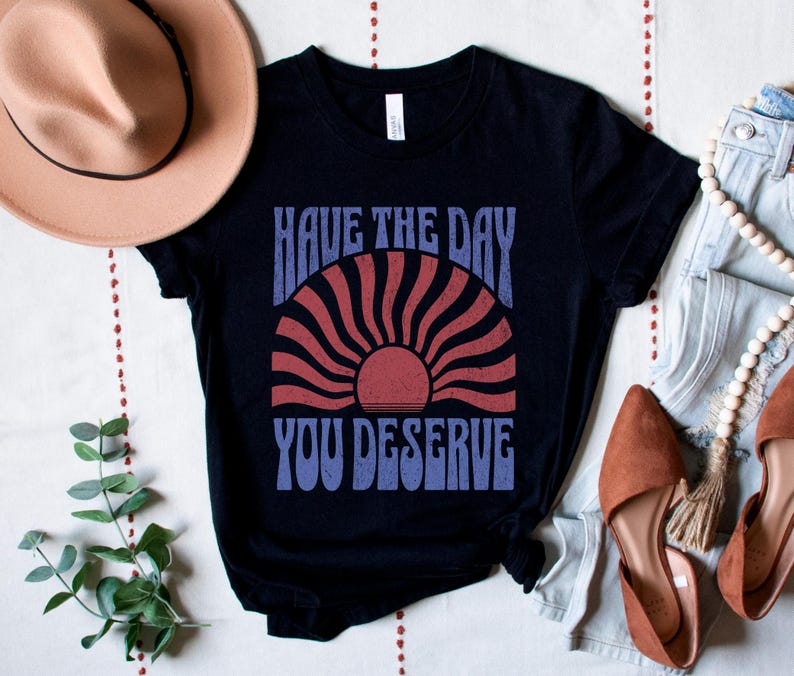 A black unisex short sleeve crew neck t-shirt that says "have the day you deserve" on the front in distressed blue retro capitalized font around a distressed image of a red wavy retro sunset