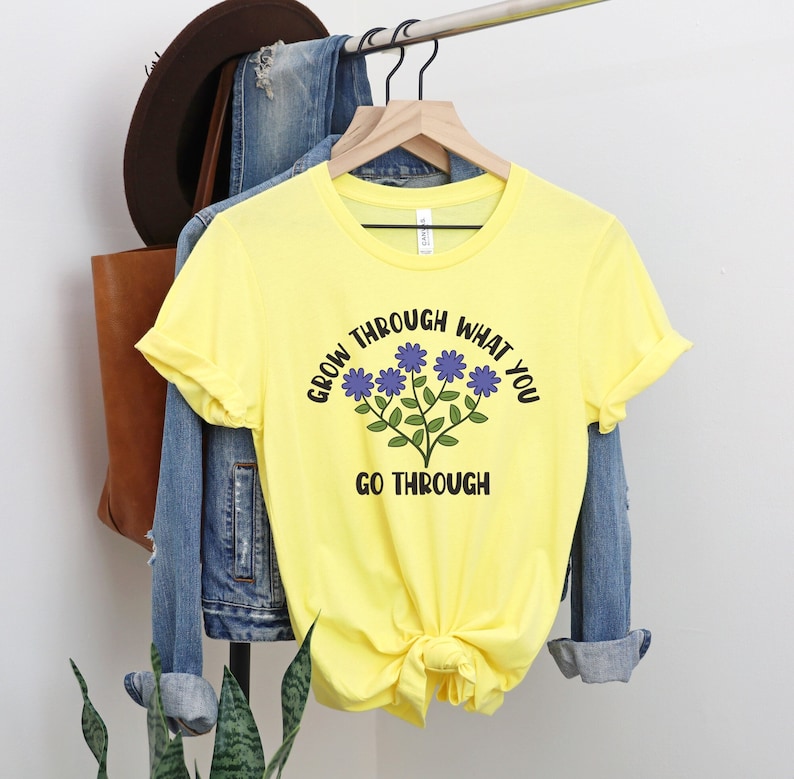 May include: A yellow t-shirt with the text "GROW THROUGH WHAT YOU GO THROUGH" in black. The shirt features a floral design with blue flowers and green stems. The shirt is hanging on a wooden hanger.