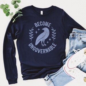 A navy blue unisex long sleeve crew neck t-shirt that says  "BECOME UNGOVERNABLE" on the chest in block text around an image of a crow, leaves, and retro stars. The whole graphic and text are light blue and have a distressed/vintage look.