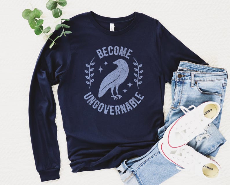 A navy blue unisex long sleeve crew neck t-shirt that says  "BECOME UNGOVERNABLE" on the chest in block text around an image of a crow, leaves, and retro stars. The whole graphic and text are light blue and have a distressed/vintage look.