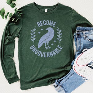 A heather forest green unisex long sleeve crew neck t-shirt that says  "BECOME UNGOVERNABLE" on the chest in block text around an image of a crow, leaves, and retro stars. The whole graphic and text are light blue and have a distressed/vintage look.