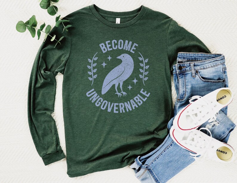 A heather forest green unisex long sleeve crew neck t-shirt that says  "BECOME UNGOVERNABLE" on the chest in block text around an image of a crow, leaves, and retro stars. The whole graphic and text are light blue and have a distressed/vintage look.