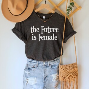 May include: A dark gray t-shirt with the text "the Future is Female" in white lettering. The shirt is hanging on a wooden hanger with a brown hat and a woven bag.