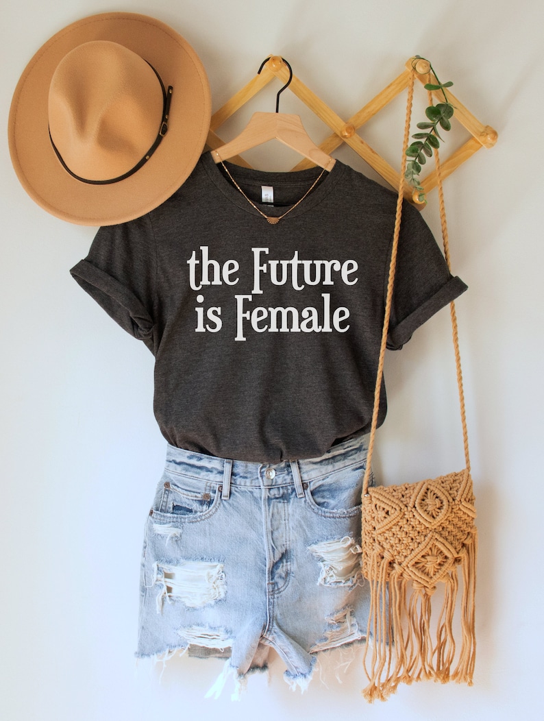May include: A dark gray t-shirt with the text "the Future is Female" in white lettering. The shirt is hanging on a wooden hanger with a brown hat and a woven bag.