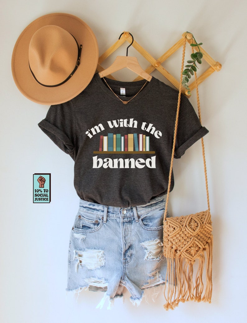 May include: A dark grey t-shirt with the text "i'm with the banned" in white. The text is stylized with a bookshelf graphic featuring colorful books. A small sticker with the text "10% to social justice" is visible in the lower left corner of the image.