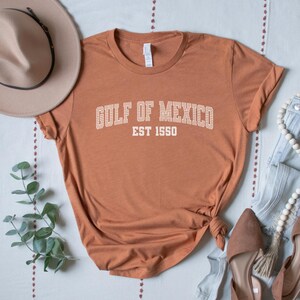 A heather autumn (dark orange) unisex crew neck short sleeve Bella+Canvas brand t-shirt that says "GULF OF MEXICO" on the chest in white outline college style font with white stripes inside the letters. The text is curved over smaller solid white letters in the same font that say "EST 1550". The graphic has a distressed/vintage look