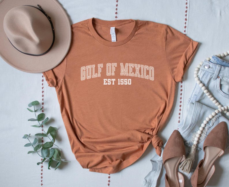 A heather autumn (dark orange) unisex crew neck short sleeve Bella+Canvas brand t-shirt that says "GULF OF MEXICO" on the chest in white outline college style font with white stripes inside the letters. The text is curved over smaller solid white letters in the same font that say "EST 1550". The graphic has a distressed/vintage look