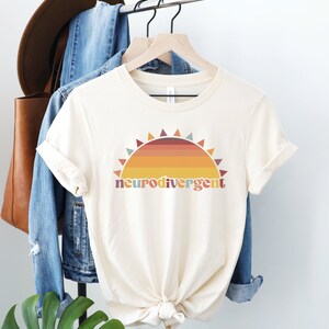 May include: A cream colored t-shirt with a retro sunset graphic and the text "neurodivergent" in colorful letters.