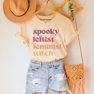 May include: A light beige t-shirt with the text "spooky leftist feminist witch" in a rainbow gradient. The shirt is on a hanger with a brown hat and a woven bag.