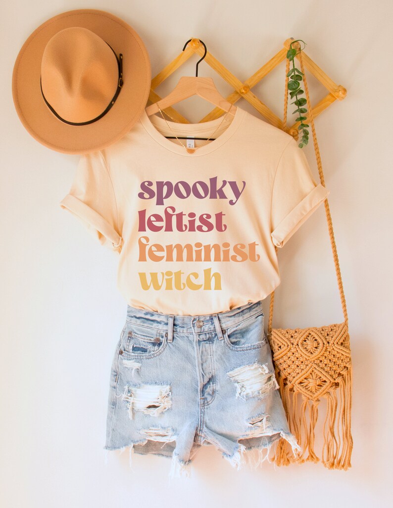 May include: A light beige t-shirt with the text "spooky leftist feminist witch" in a rainbow gradient. The shirt is on a hanger with a brown hat and a woven bag.