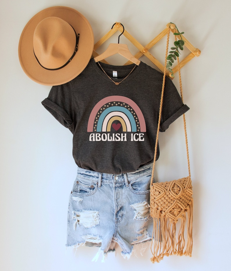 May include: A dark grey t-shirt with a rainbow graphic and the text "ABOLISH ICE" in white. The rainbow has a red heart in the center and stars around it. The shirt is hanging on a wooden hanger with a brown hat and a woven bag.