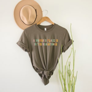 May include: A brown t-shirt with the text "A woman's place is in the resistance" printed in colourful letters.