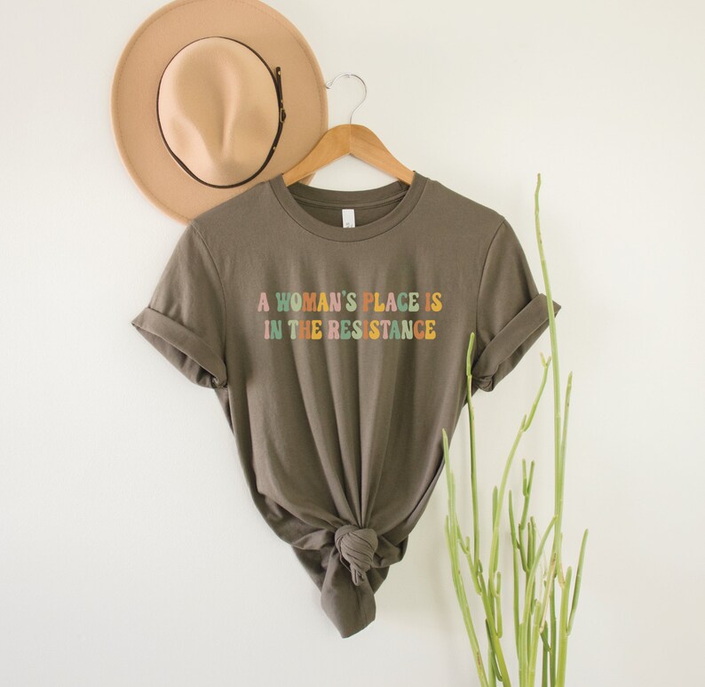 May include: A brown t-shirt with the text "A woman's place is in the resistance" printed in colourful letters.