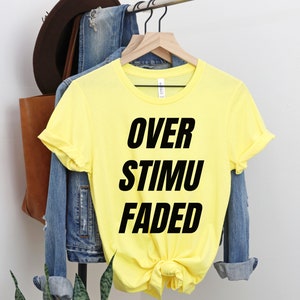 May include: A yellow t-shirt with the words "OVER STIMU FADED" printed in bold black letters. The shirt is hanging on a wooden hanger, with a denim jacket and a brown leather bag in the background.