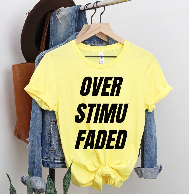 May include: A yellow t-shirt with the words "OVER STIMU FADED" printed in bold black letters. The shirt is hanging on a wooden hanger, with a denim jacket and a brown leather bag in the background.