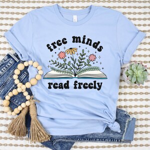 A baby blue unisex crew neck short sleeve t-shirt with black text saying "free minds read freely" around a graphic of a book with colorful flowers and stars coming out of it