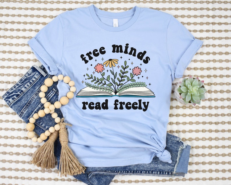 A baby blue unisex crew neck short sleeve t-shirt with black text saying "free minds read freely" around a graphic of a book with colorful flowers and stars coming out of it