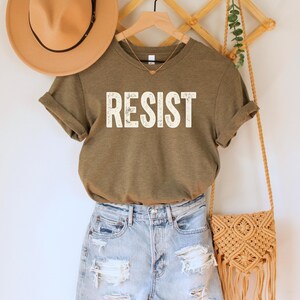 A heather olive green/brown unisex crew neck short sleeve Bella+Canvas t-shirt that says "RESIST" in distressed white tall block text