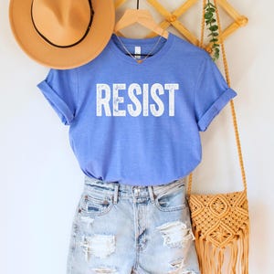 A heather Columbia (medium) blue unisex crew neck short sleeve Bella+Canvas t-shirt that says "RESIST" in distressed white tall block text