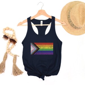 May include: A navy blue racerback tank top with a distressed, rainbow pride flag design. The flag includes the colors of the traditional rainbow flag, as well as the colors of the transgender pride flag.
