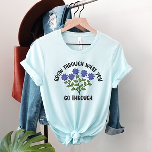 May include: A light blue t-shirt with the text "GROW THROUGH WHAT YOU GO THROUGH" in black and a floral design in purple and green. The shirt is tied at the waist and hangs on a wooden hanger.