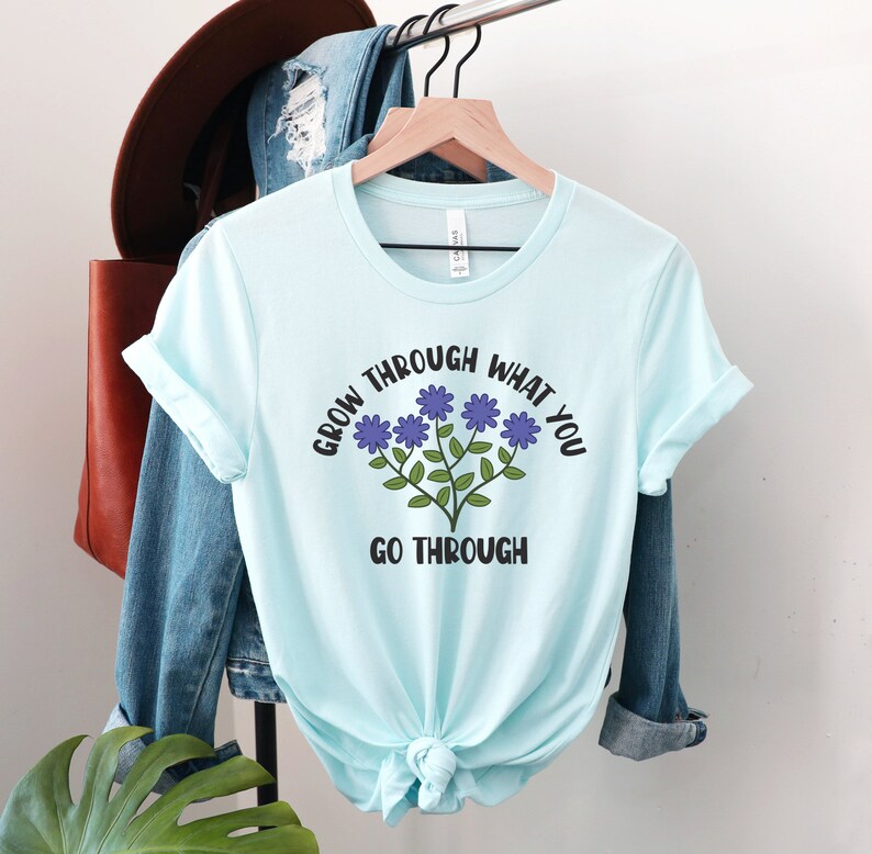 May include: A light blue t-shirt with the text "GROW THROUGH WHAT YOU GO THROUGH" in black and a floral design in purple and green. The shirt is tied at the waist and hangs on a wooden hanger.