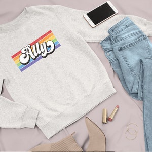 Pu&ograve; includere: Una felpa grigia con un design a righe arcobaleno e la parola "Ally" in un font retr&ograve;.