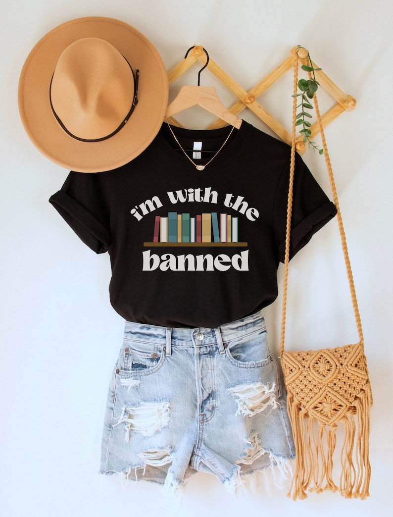 May include: Black t-shirt with the text "i'm with the banned" in white with a graphic of a bookshelf with colorful books. The shirt is on a hanger with a brown hat and a woven bag.
