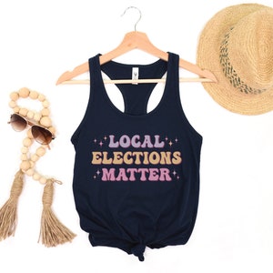 A navy blue women’s racerback tank top that says "local elections matter" in purple, yellow, and pink retro text with retro stars
