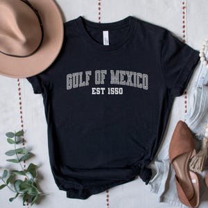 A black unisex crew neck short sleeve Bella+Canvas brand t-shirt that says "GULF OF MEXICO" on the chest in white outline college style font with white stripes inside the letters. The text is curved over smaller solid white letters in the same font that say "EST 1550". The graphic has a distressed/vintage look