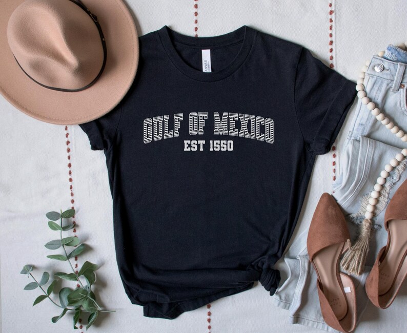 A black unisex crew neck short sleeve Bella+Canvas brand t-shirt that says "GULF OF MEXICO" on the chest in white outline college style font with white stripes inside the letters. The text is curved over smaller solid white letters in the same font that say "EST 1550". The graphic has a distressed/vintage look
