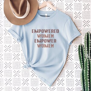 May include: A light blue t-shirt with the text "EMPOWERED WOMEN EMPOWER WOMEN" printed in brown on the front. The shirt is hanging on a wooden hanger against a white background with a black and white geometric pattern.