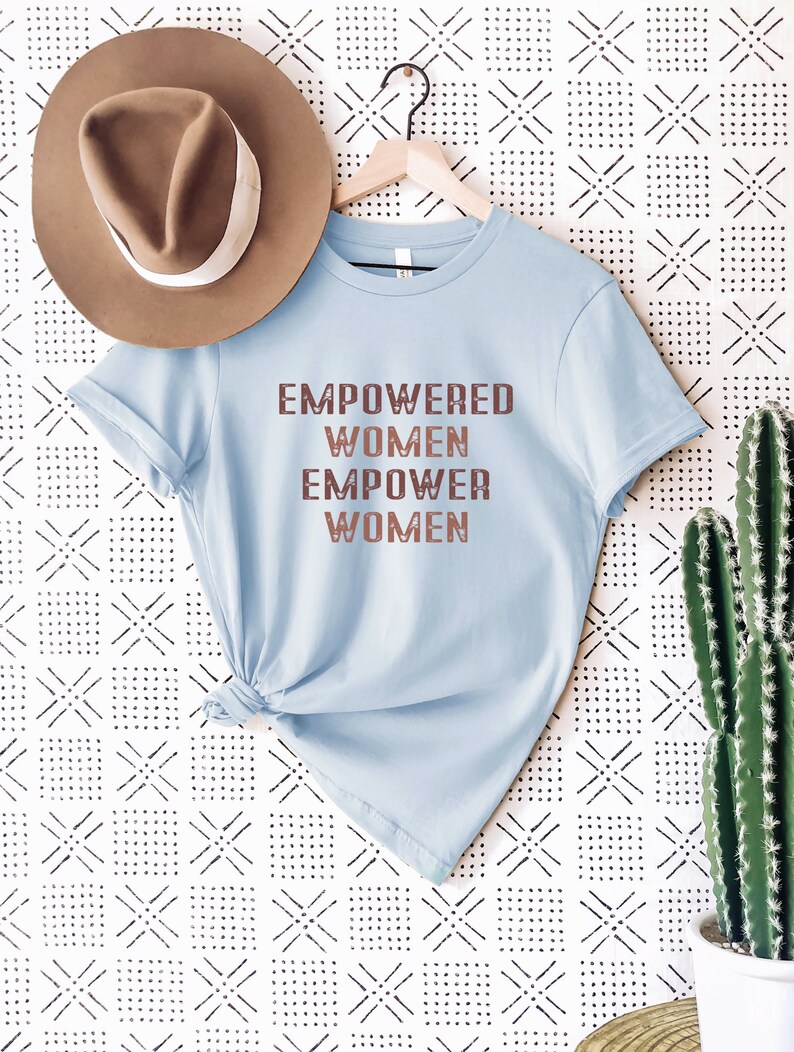 May include: A light blue t-shirt with the text "EMPOWERED WOMEN EMPOWER WOMEN" printed in brown on the front. The shirt is hanging on a wooden hanger against a white background with a black and white geometric pattern.