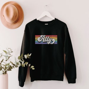 Pu&ograve; includere: Felpa nera con un design a righe arcobaleno e la parola "Ally" in un font retr&ograve;.