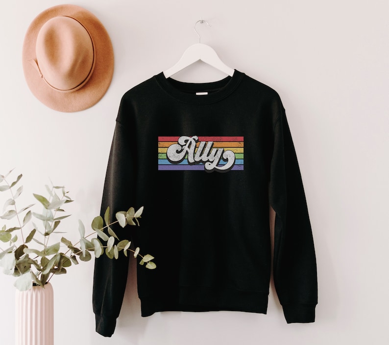 Pu&ograve; includere: Felpa nera con un design a righe arcobaleno e la parola "Ally" in un font retr&ograve;.