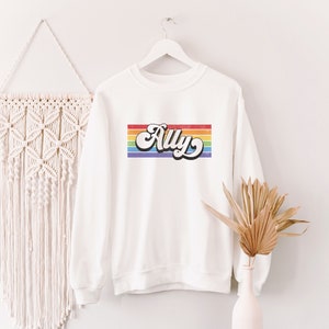 Pu&ograve; includere: Felpa bianca con un design a righe arcobaleno e il nome "Ally" in un font retr&ograve;.