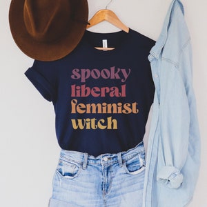 A navy unisex crew neck t-shirt with the phrase "spooky liberal feminist witch" in purple, red, orange, and yellow text