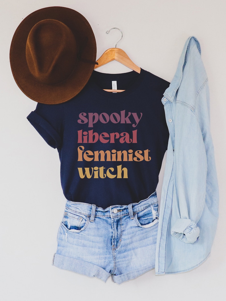 A navy unisex crew neck t-shirt with the phrase "spooky liberal feminist witch" in purple, red, orange, and yellow text