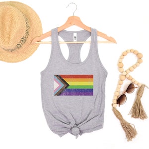 May include: A gray tank top with a vintage-style, multi-colored pride flag printed on the front. The flag includes the colors of the transgender pride flag, the black and brown stripes of the progress pride flag, and the traditional rainbow pride flag.