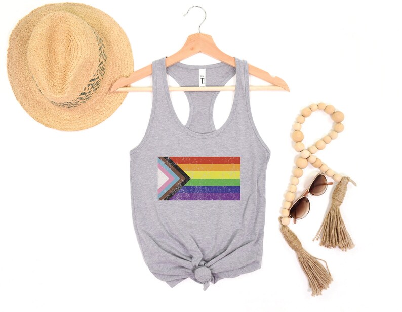 May include: A gray tank top with a vintage-style, multi-colored pride flag printed on the front. The flag includes the colors of the transgender pride flag, the black and brown stripes of the progress pride flag, and the traditional rainbow pride flag.