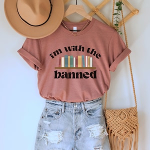 May include: A mauve t-shirt with the text "i'm with the banned" and a graphic of a bookshelf with colorful books. The shirt is on a hanger with a brown hat and a woven bag.