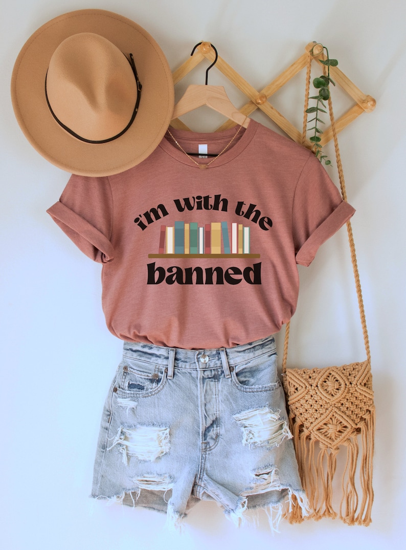 May include: A mauve t-shirt with the text "i'm with the banned" and a graphic of a bookshelf with colorful books. The shirt is on a hanger with a brown hat and a woven bag.