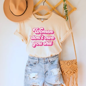 A natural (light beige) unisex crew neck t-shirt with the words "women don't owe you shit" in retro pink text with retro pink stars