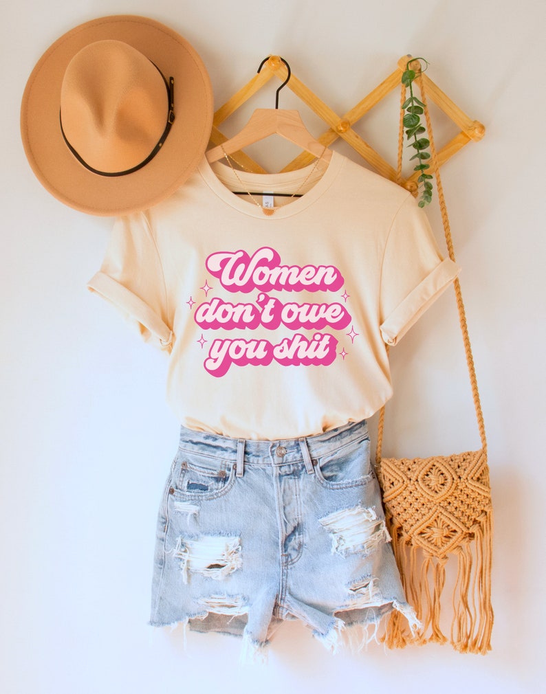 A natural (light beige) unisex crew neck t-shirt with the words "women don't owe you shit" in retro pink text with retro pink stars