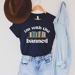 May include: A navy blue t-shirt with the text "i'm with the banned" and a graphic of a bookshelf with colorful books. The shirt is hanging on a wooden hanger.