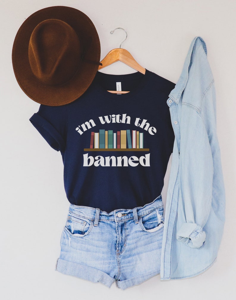 May include: A navy blue t-shirt with the text "i'm with the banned" and a graphic of a bookshelf with colorful books. The shirt is hanging on a wooden hanger.