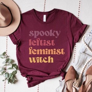 May include: A maroon t-shirt with the text "spooky leftist feminist witch" in a gradient of colors from pink to orange.