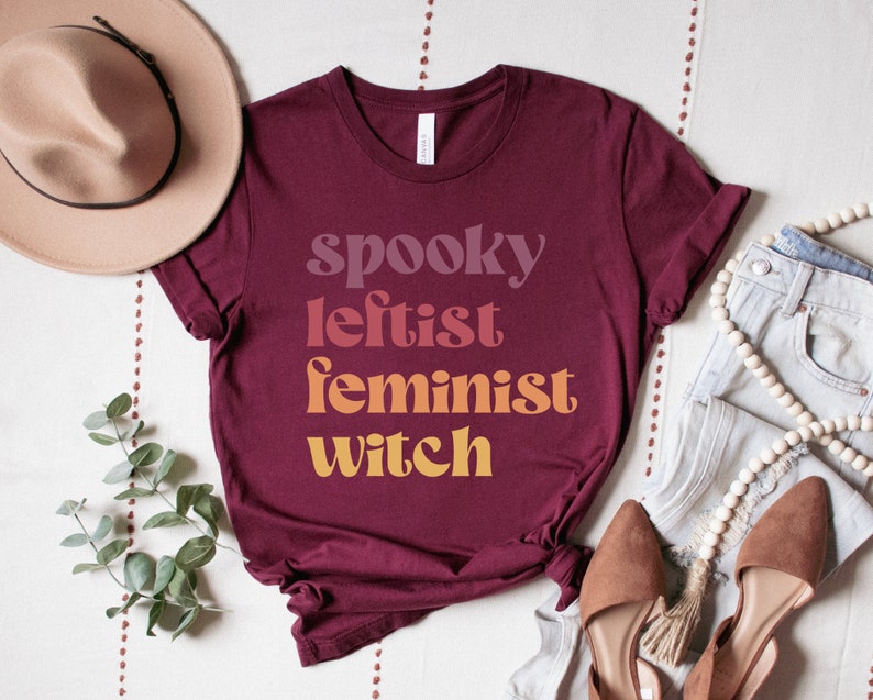 May include: A maroon t-shirt with the text "spooky leftist feminist witch" in a gradient of colors from pink to orange.